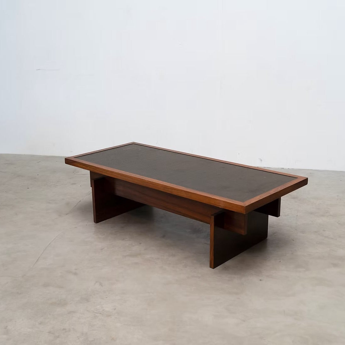 A mid-century modern wooden coffee table with a dark brown leather top and angled wooden legs, set against a plain white wall and concrete floor.