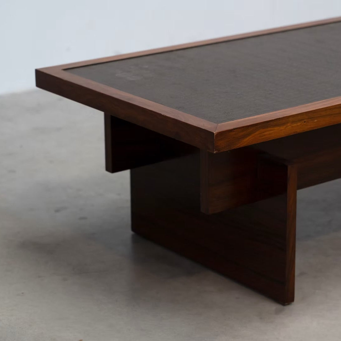 A mid-century modern wooden coffee table with a dark brown leather top and a dark wood frame, shown in a minimalist room setting with a concrete floor.