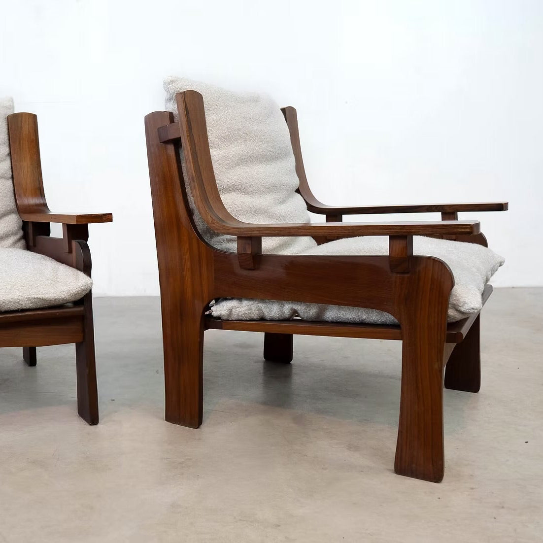 Two mid-century modern wooden armchairs with curved dark wood frames and light beige textured cushions, shown on a concrete floor against a white wall.