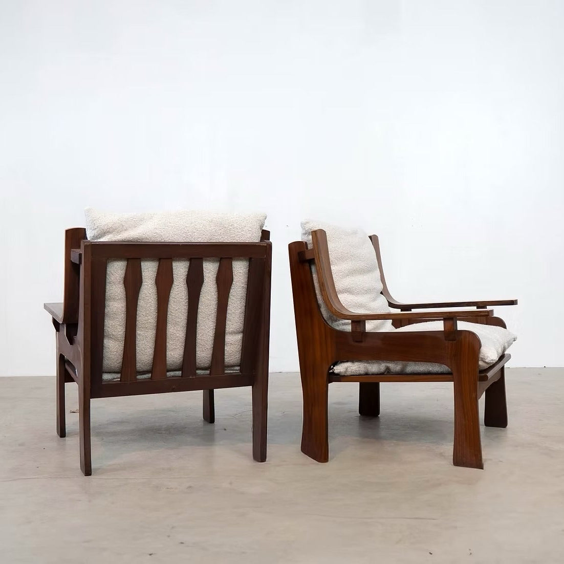 Two mid-century modern wooden armchairs with cream cushions, shown from the back in a minimalist studio setting with a white wall and concrete floor.