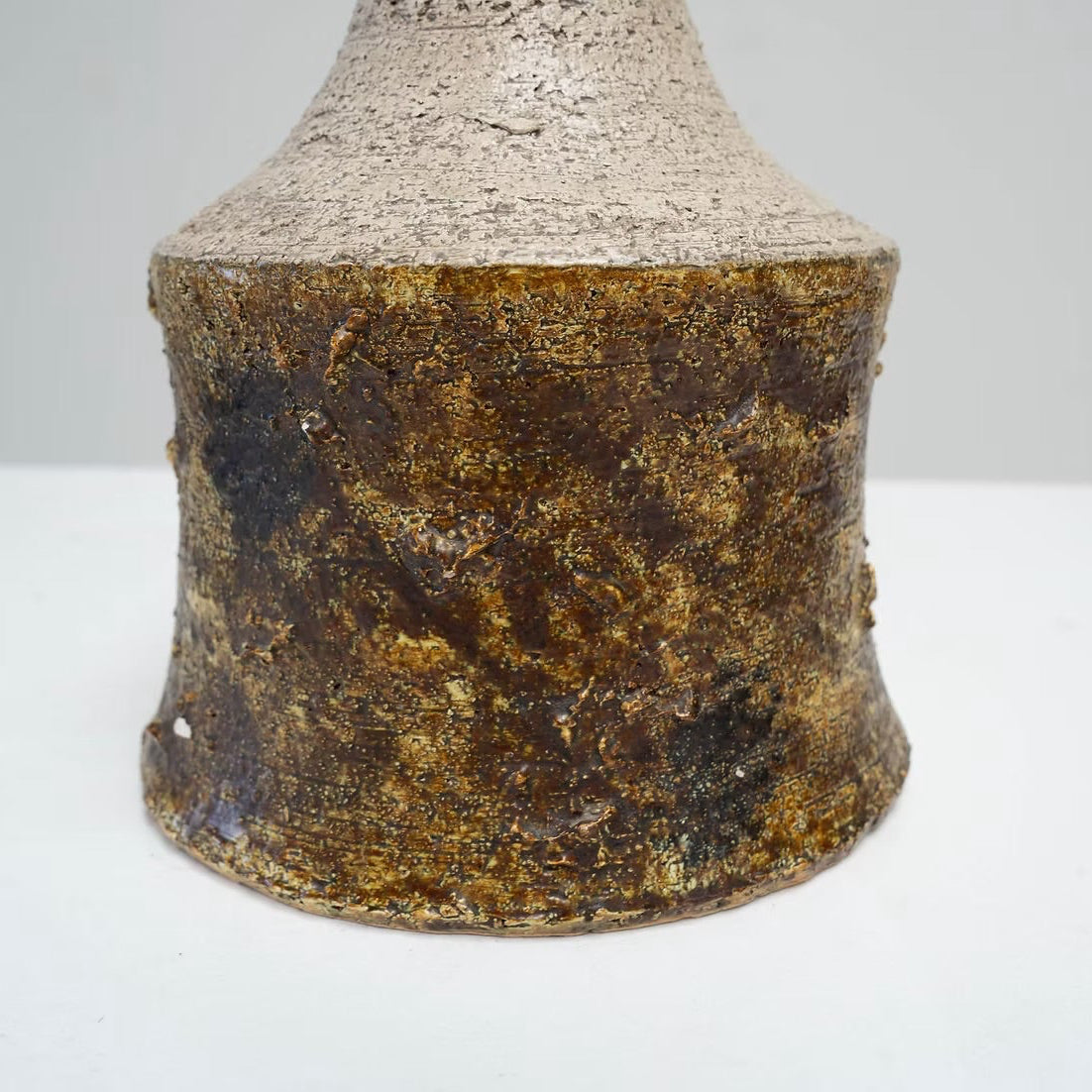 A close-up of a textured ceramic vase or bottle base with a mottled metallic gold and dark brown glaze, standing on a white surface against a light gray background.