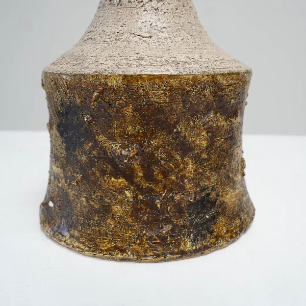 A close-up of a textured ceramic vase or bottle base with a mottled metallic gold and dark brown glaze, standing on a white surface against a light gray background.