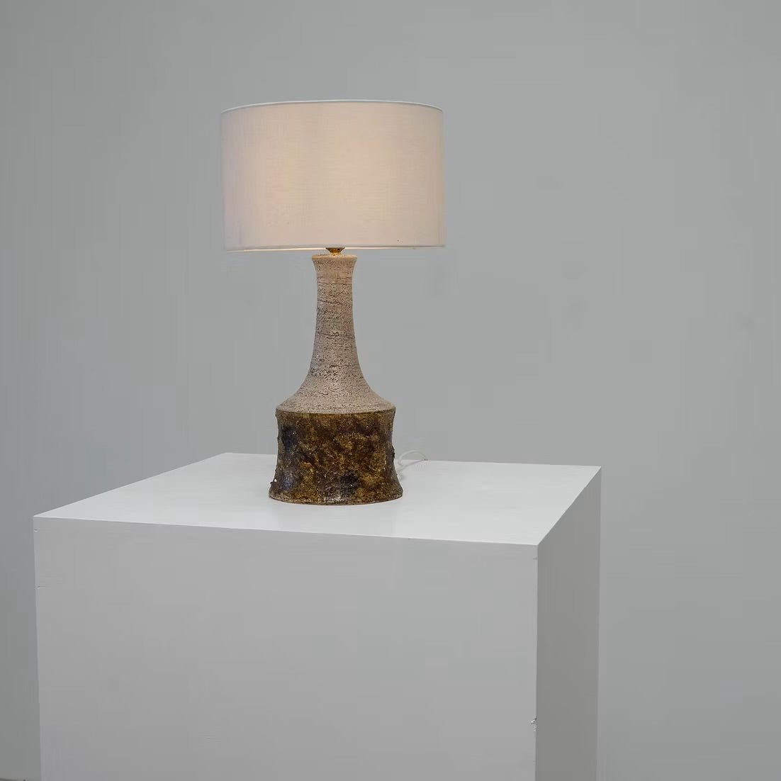 A table lamp with a textured beige and brown ceramic base and a white drum shade, displayed on a white pedestal against a light gray wall.