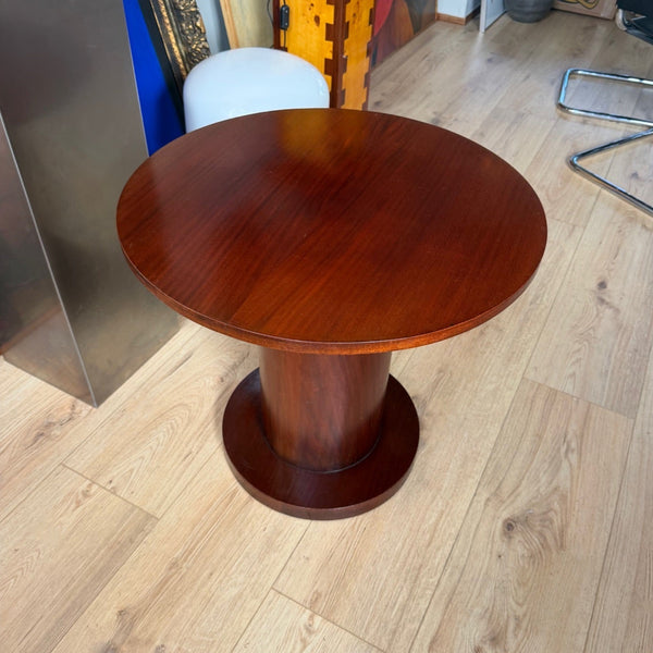 A round wooden side table with a polished reddish-brown finish and a pedestal base, shown in a showroom with light wood floors and framed art in the background.