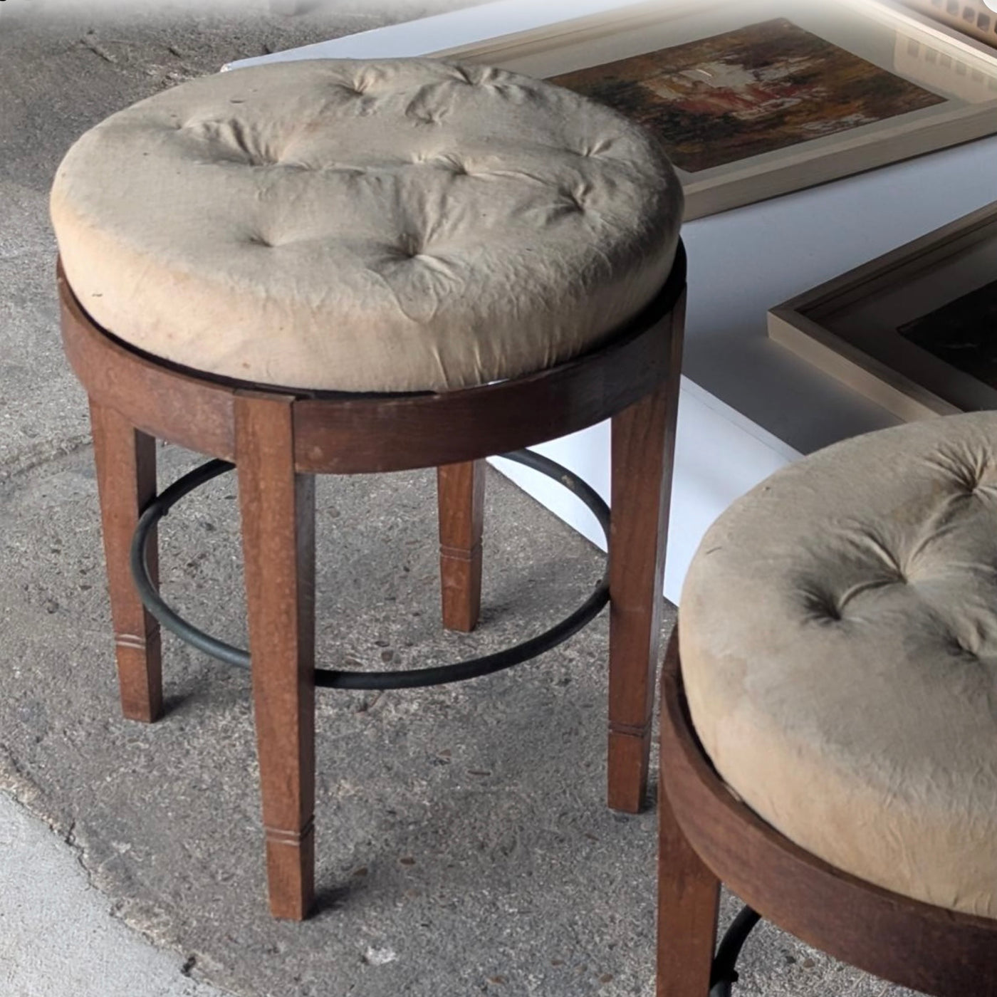 A vintage-style bar stool with a round tufted beige fabric cushion on a dark brown wooden frame, set on a concrete floor next to another matching stool.