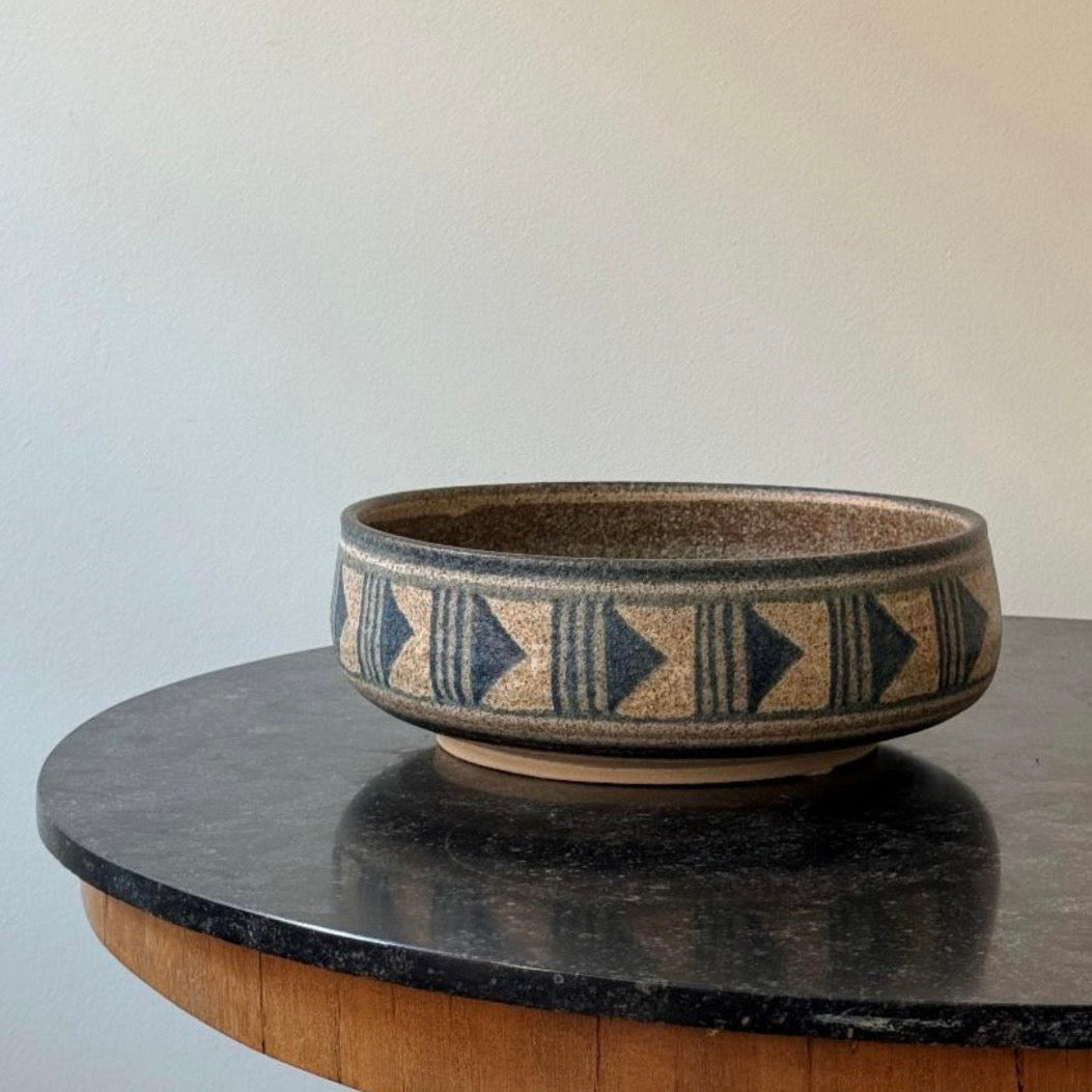 A round ceramic bowl with a textured beige and brown glaze, decorated with a dark blue geometric triangle pattern around the outer rim, displayed on a dark stone tabletop against a plain wall.