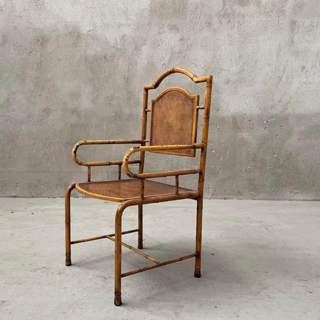 A vintage-style armchair made of natural bamboo with a woven cane seat and backrest, set against a textured gray concrete wall and floor.
