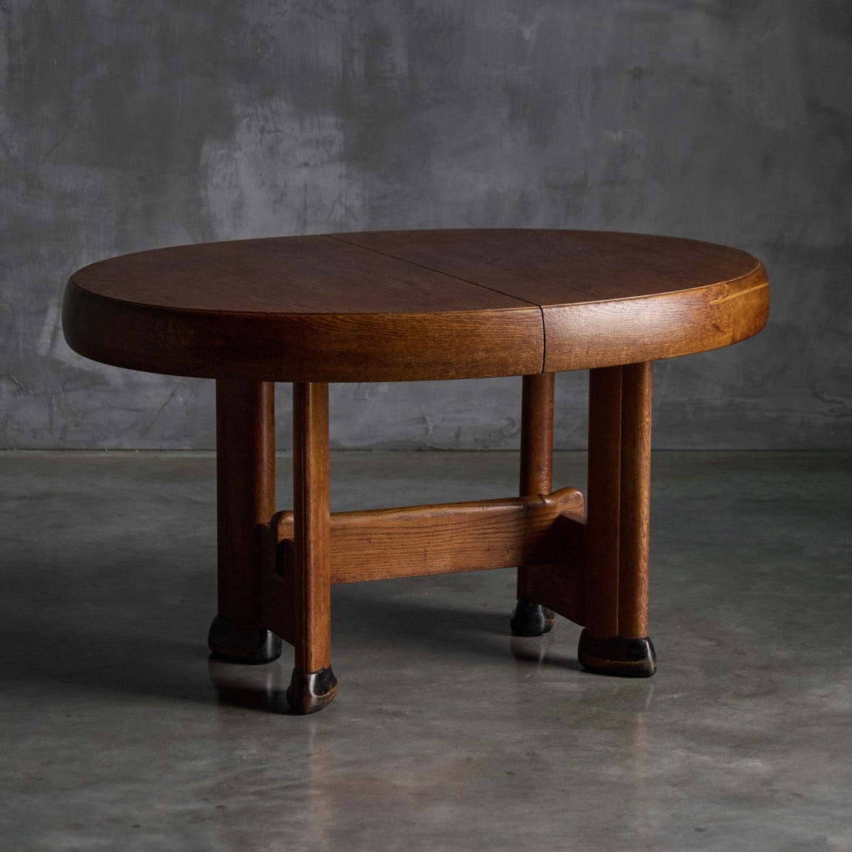 A round wooden table with a warm medium-brown finish and dark feet, set against a textured gray concrete wall and floor.
