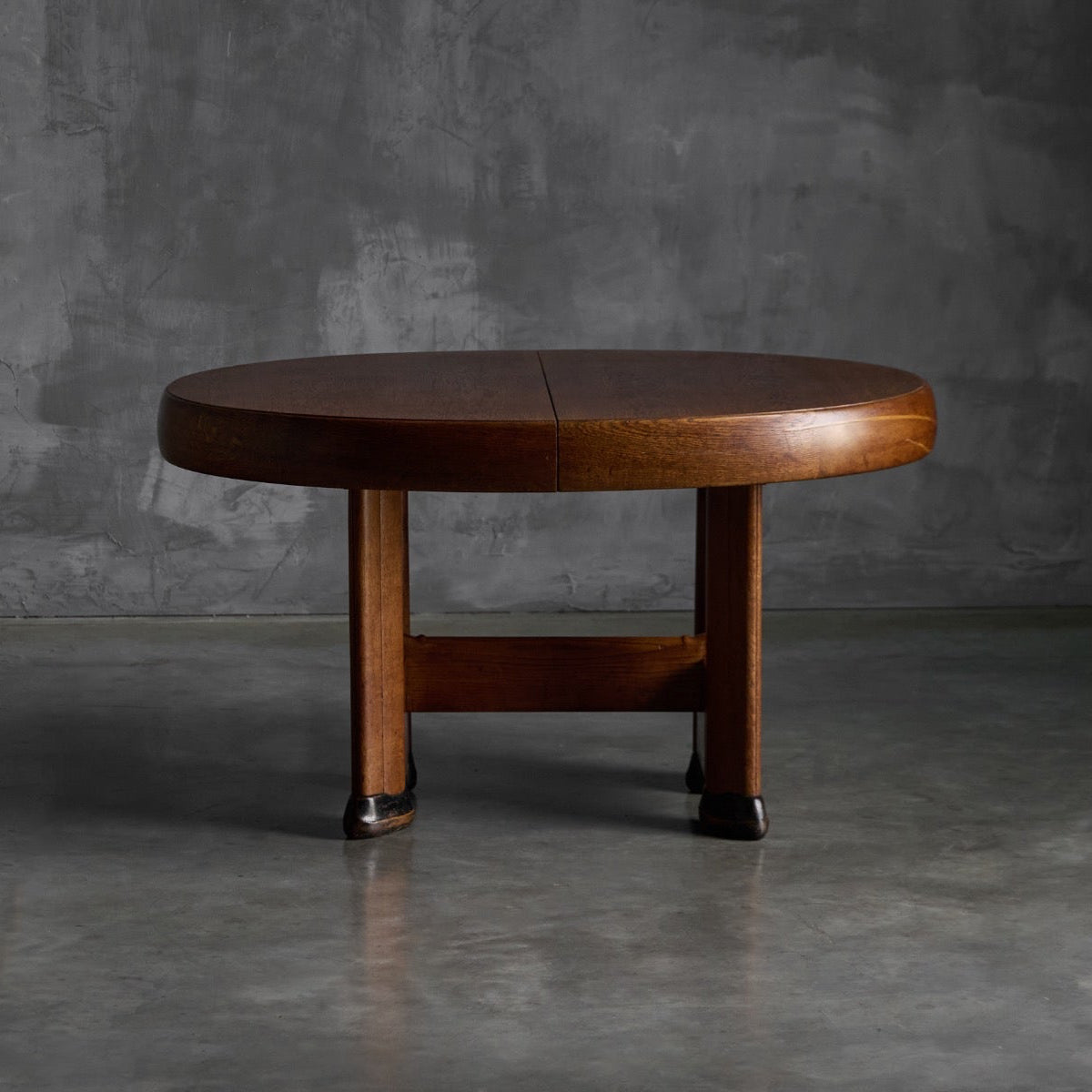 A round wooden dining table  photographed in a minimalist studio setting with a textured gray concrete wall and floor.