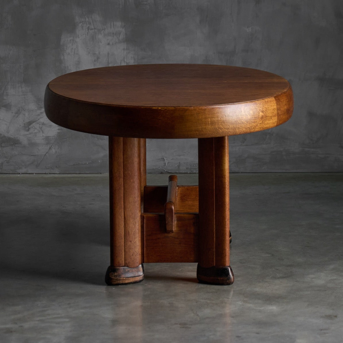 A round wooden stool with a thick circular seat and four sturdy legs, shown in a studio setting against a textured gray background.
