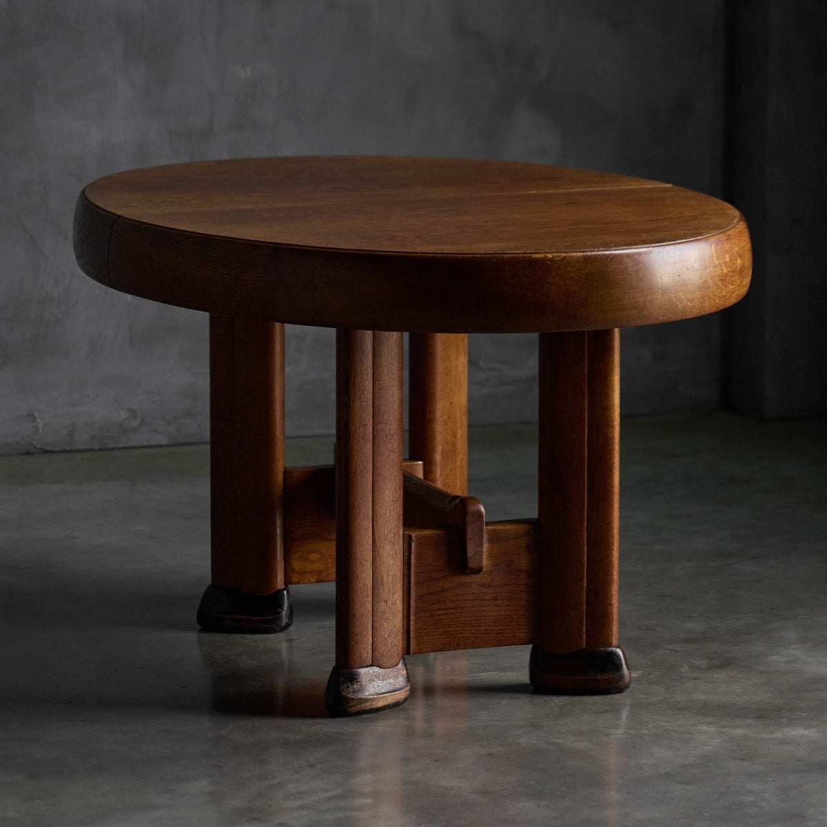 A round wooden stool with a smooth, polished top and four sturdy legs, set against a dark gray concrete wall and floor.