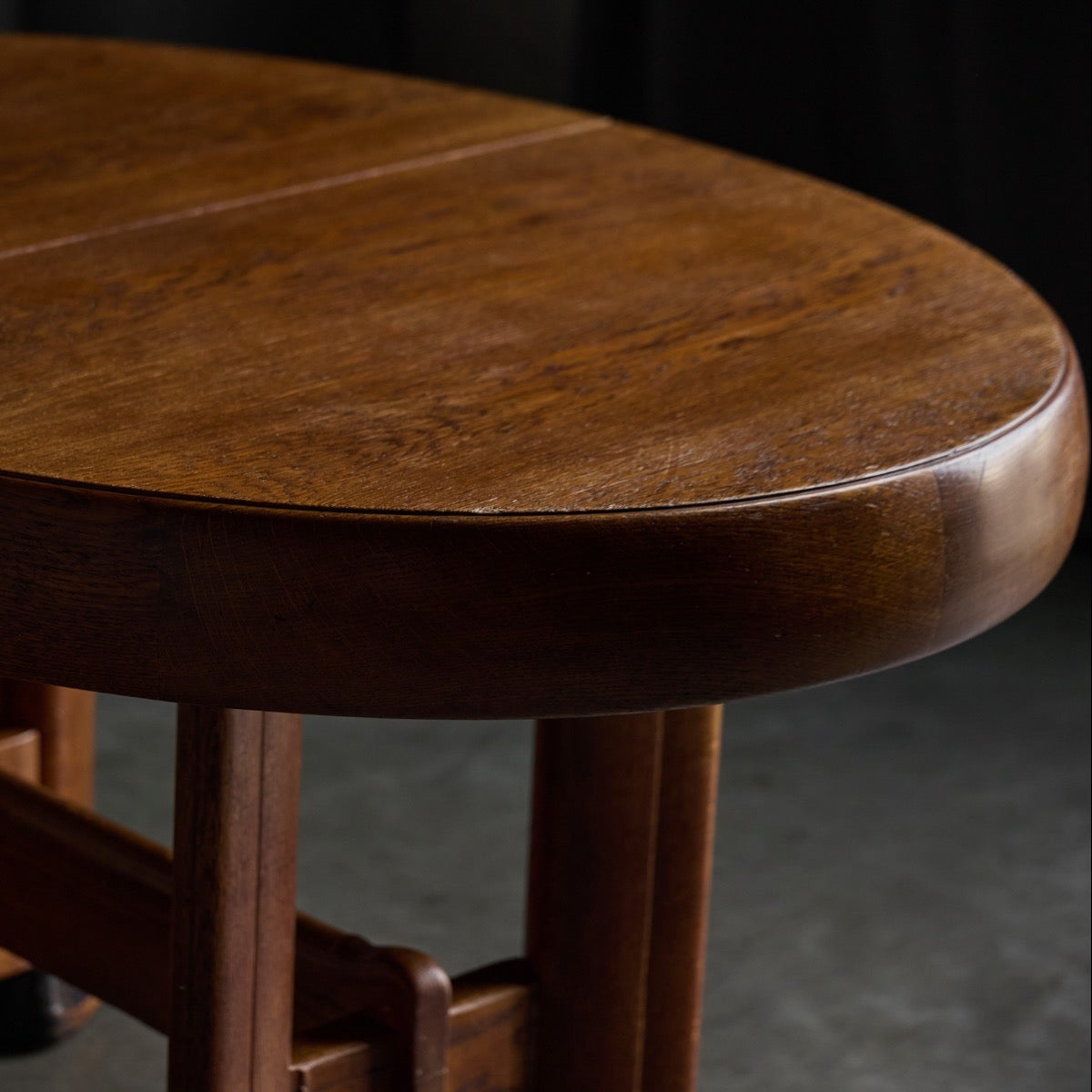 Close-up of an oval wooden table with a rich medium-brown finish, showing the tabletop and part of the support legs against a dark background.