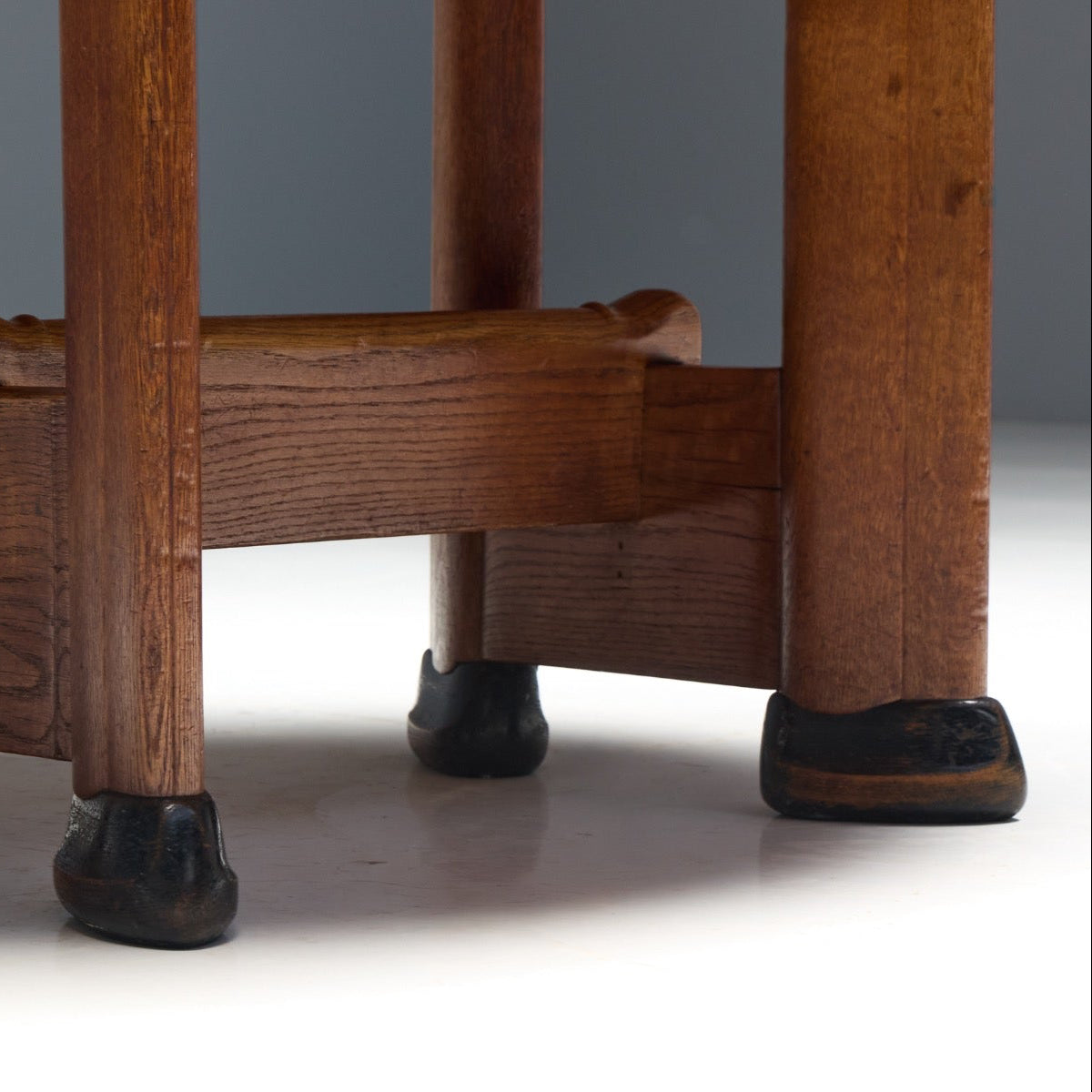 Close-up of the lower portion of a wooden stool or table leg frame, showing four dark-stained legs with black feet and a central wooden support structure on a white floor against a gray background.