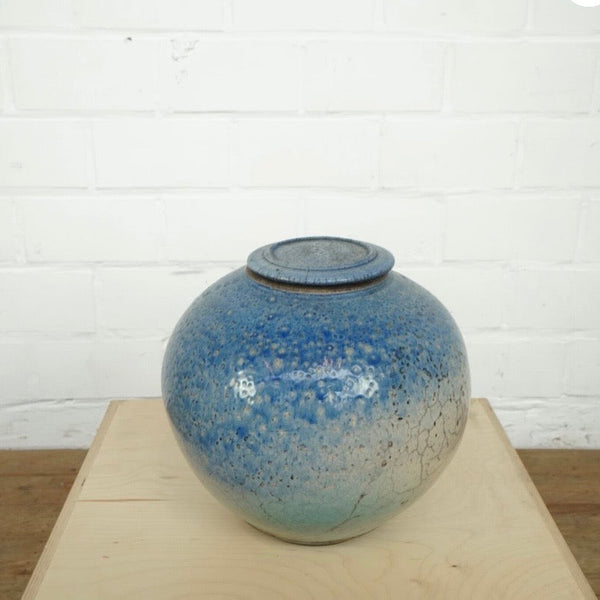 A round glazed ceramic lidded jar in shades of blue and teal, displayed on a light wood block against a white brick wall background.