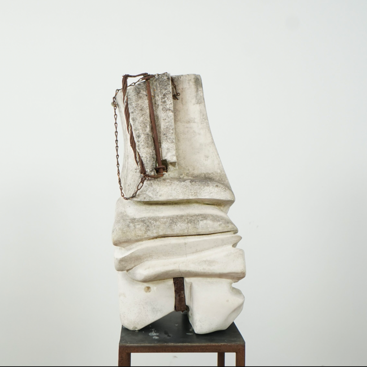 An abstract sculptural artwork made of stacked off-white stone or concrete forms with a dark brown chain-like element, displayed on a small dark pedestal against a plain light background.