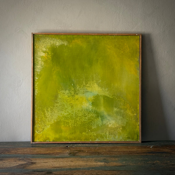 A framed abstract painting featuring various shades of green with a textured surface, displayed on a wooden floor against a white wall.