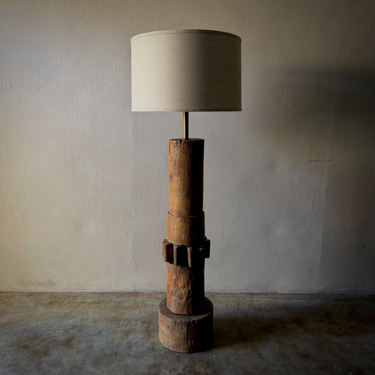 A rustic industrial-style floor lamp featuring a stacked natural wood base with a dark metal band detail, topped with a slim brass stem and a large cylindrical beige fabric drum shade, set against a textured plaster wall and concrete floor.