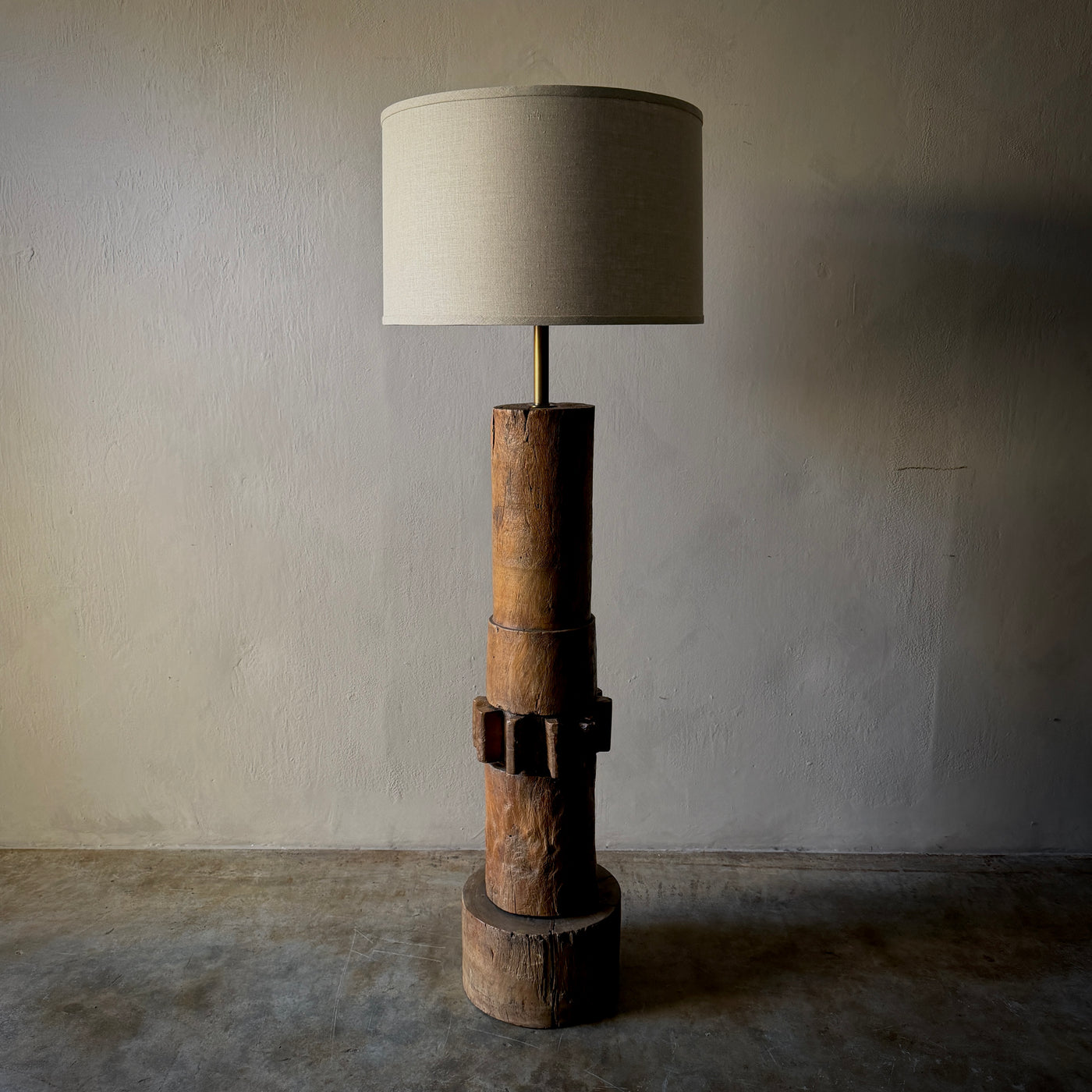 A rustic industrial-style floor lamp featuring a stacked natural wood base with a dark metal band detail, topped with a slim brass stem and a large cylindrical beige fabric drum shade, set against a textured plaster wall and concrete floor.