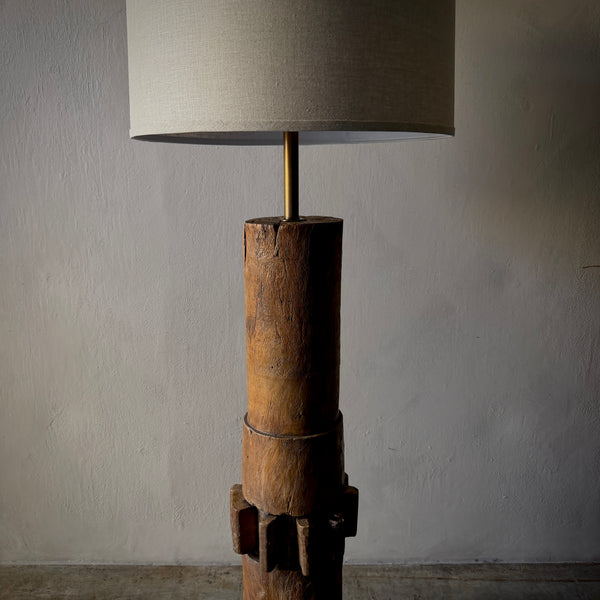 A rustic wooden table lamp with a cylindrical carved wood base and a beige fabric drum shade, set against a neutral textured wall.