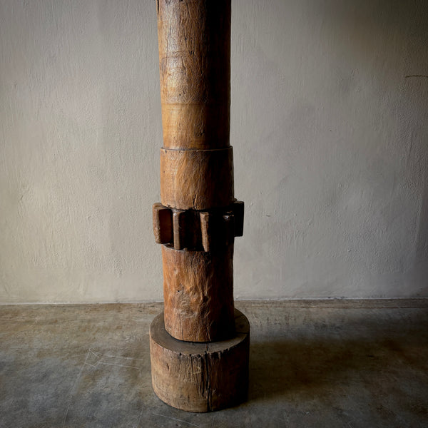 A rustic wooden sculpture or furniture leg featuring a cylindrical post with a textured, weathered finish and a dark metal band with protruding tabs, mounted on a round wooden base against a neutral wall.