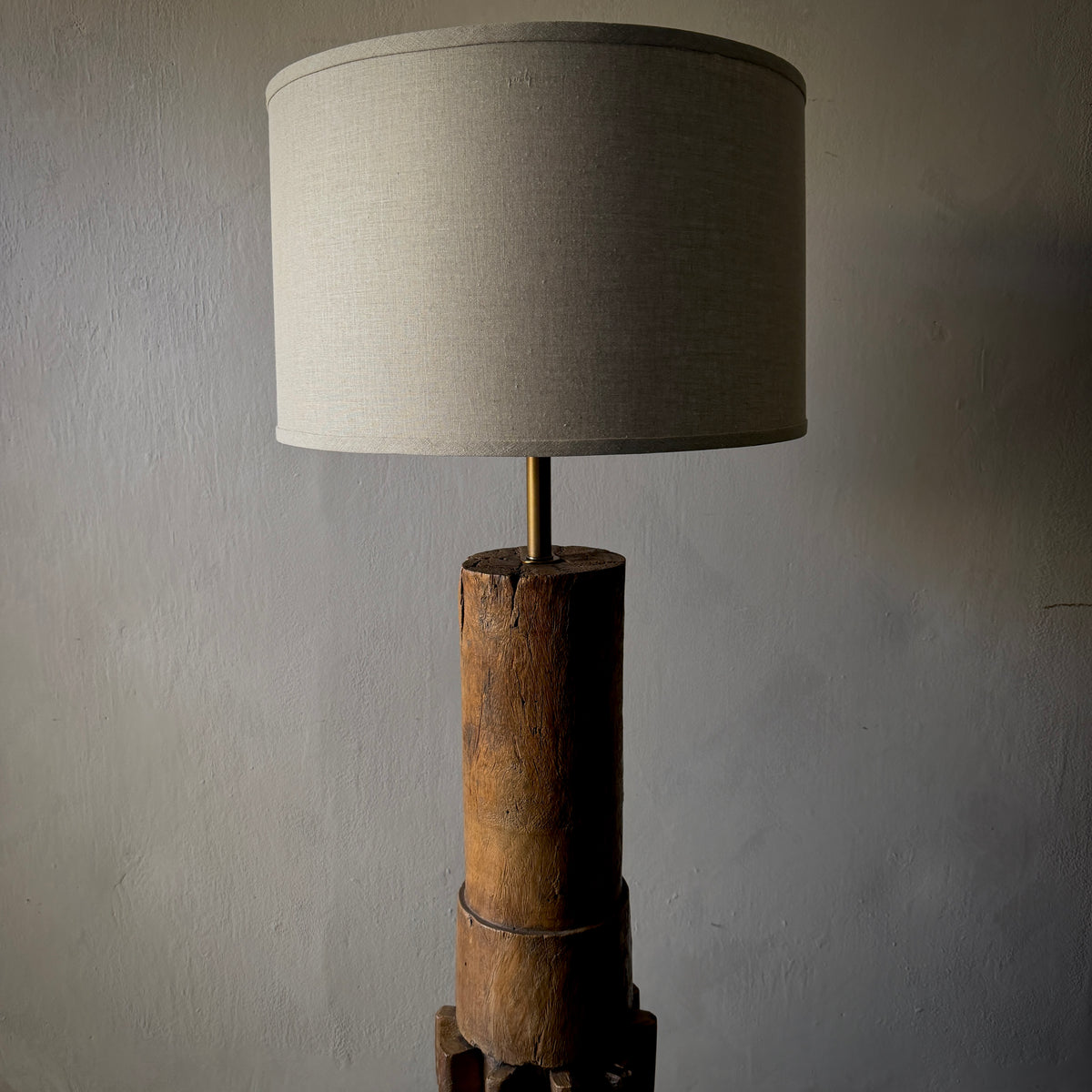 A rustic table lamp featuring a natural wood cylindrical base and a light beige linen drum shade, set against a textured gray wall.