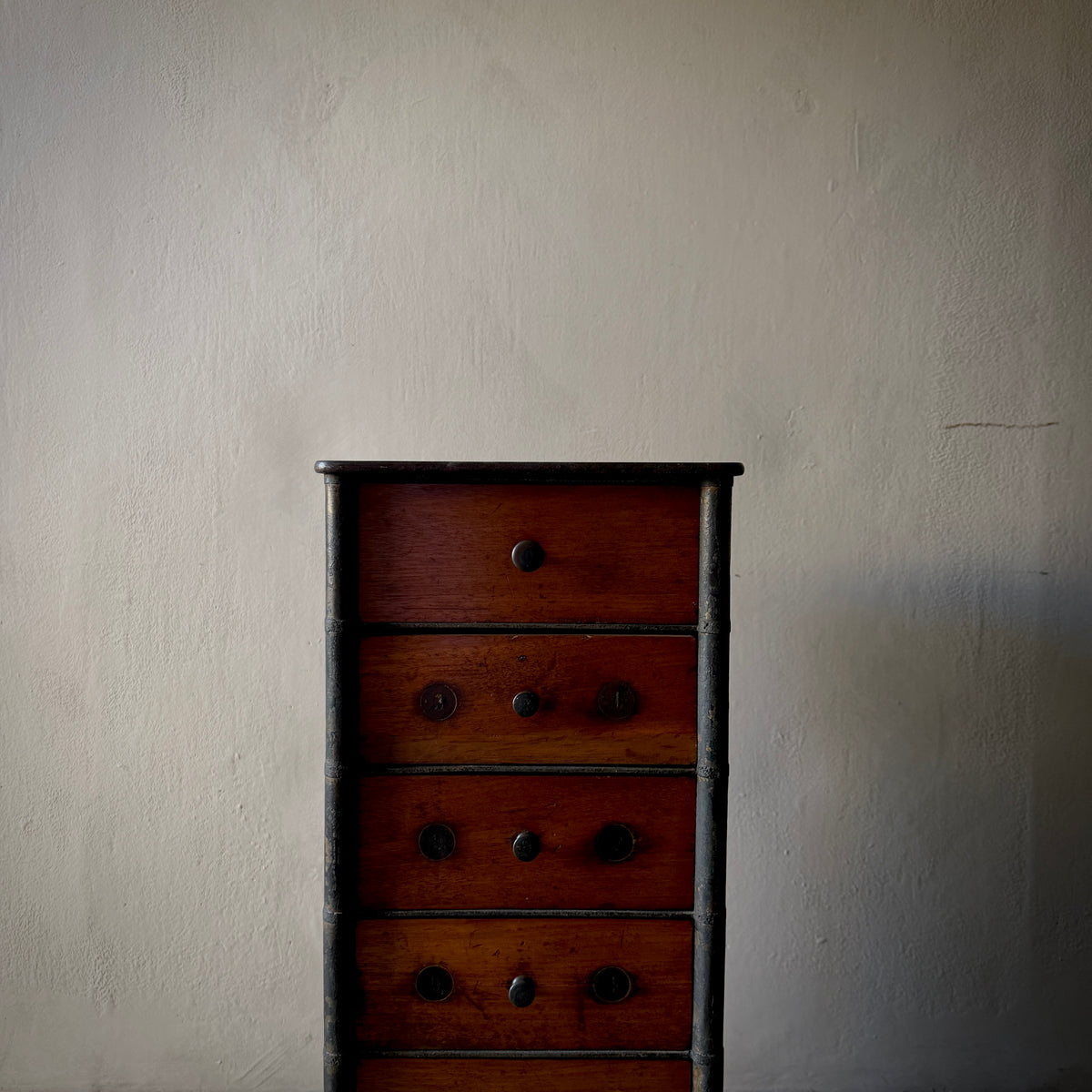 A vintage wooden chest of drawers with five drawers and dark metal knobs, set against a textured off-white wall.