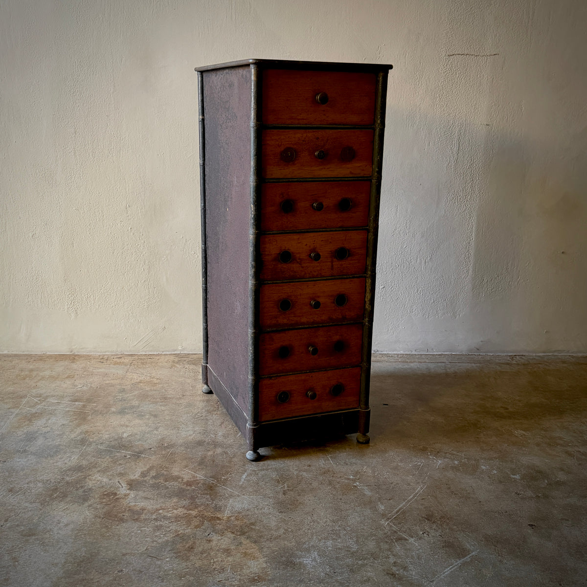 A vintage seven-drawer chest of drawers with reddish-brown wood fronts and dark metal frames, standing on casters in a room with a textured beige wall and concrete floor.