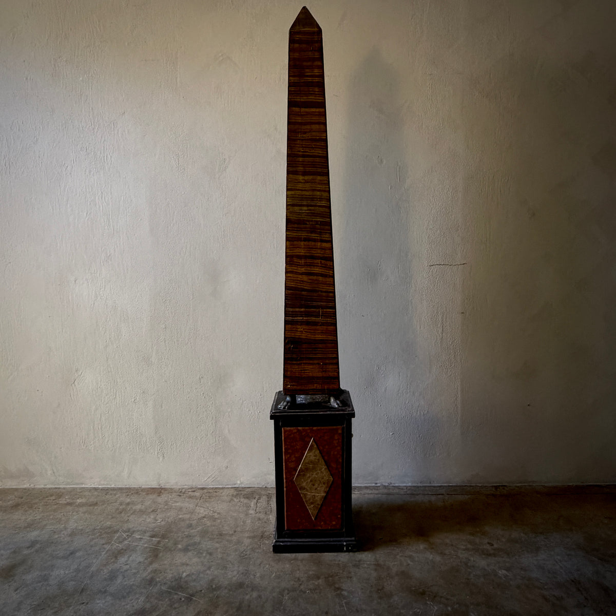 A tall Egyptian obelisk sculpture with a dark brown striped wood body and a black base featuring a gold diamond motif, set against a textured beige wall and concrete floor.