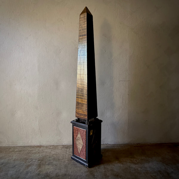 A tall wooden obelisk sculpture with a tapered, pointed top, mounted on a decorative rectangular base with a geometric mosaic pattern, set against a textured beige wall and concrete floor.