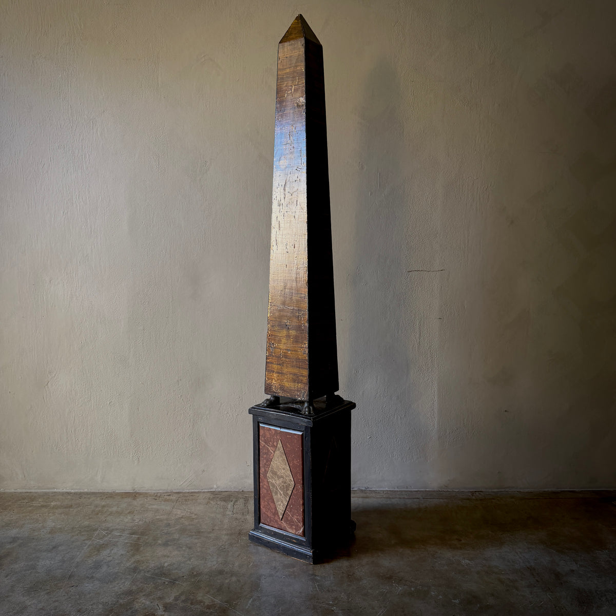 A tall wooden obelisk sculpture with a dark base, standing against a plain beige wall on a concrete floor.