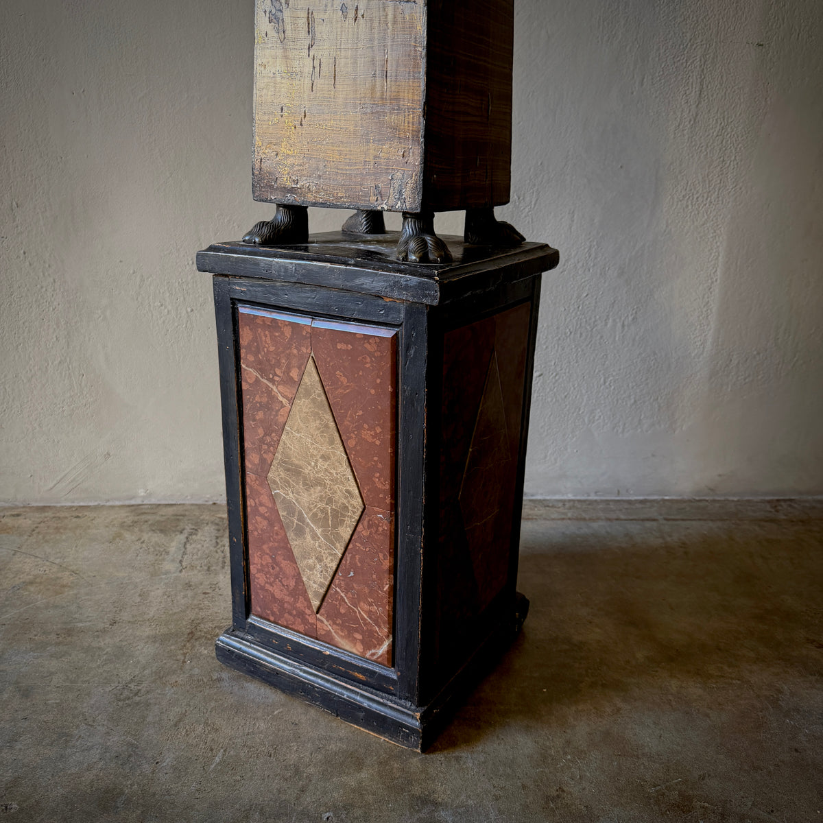 A vintage industrial pedestal lamp base with a black painted wood frame, reddish-brown marbled front panels, and a weathered wooden upper section, set against a plain concrete floor and wall.