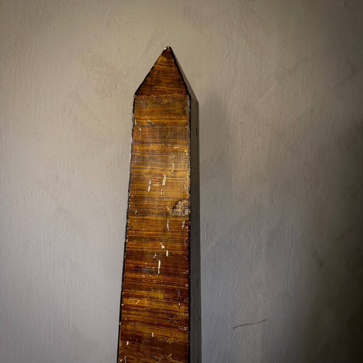 A tall, narrow wooden wall-mounted obelisk or decorative board with a pointed top, shown against a plain textured wall.