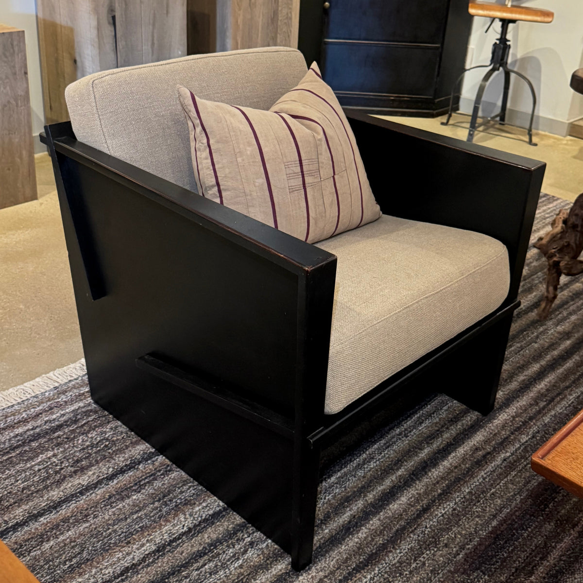 A modern accent chair with a black wooden frame and light beige upholstered seat and back, styled with a striped throw pillow in an indoor showroom setting.
