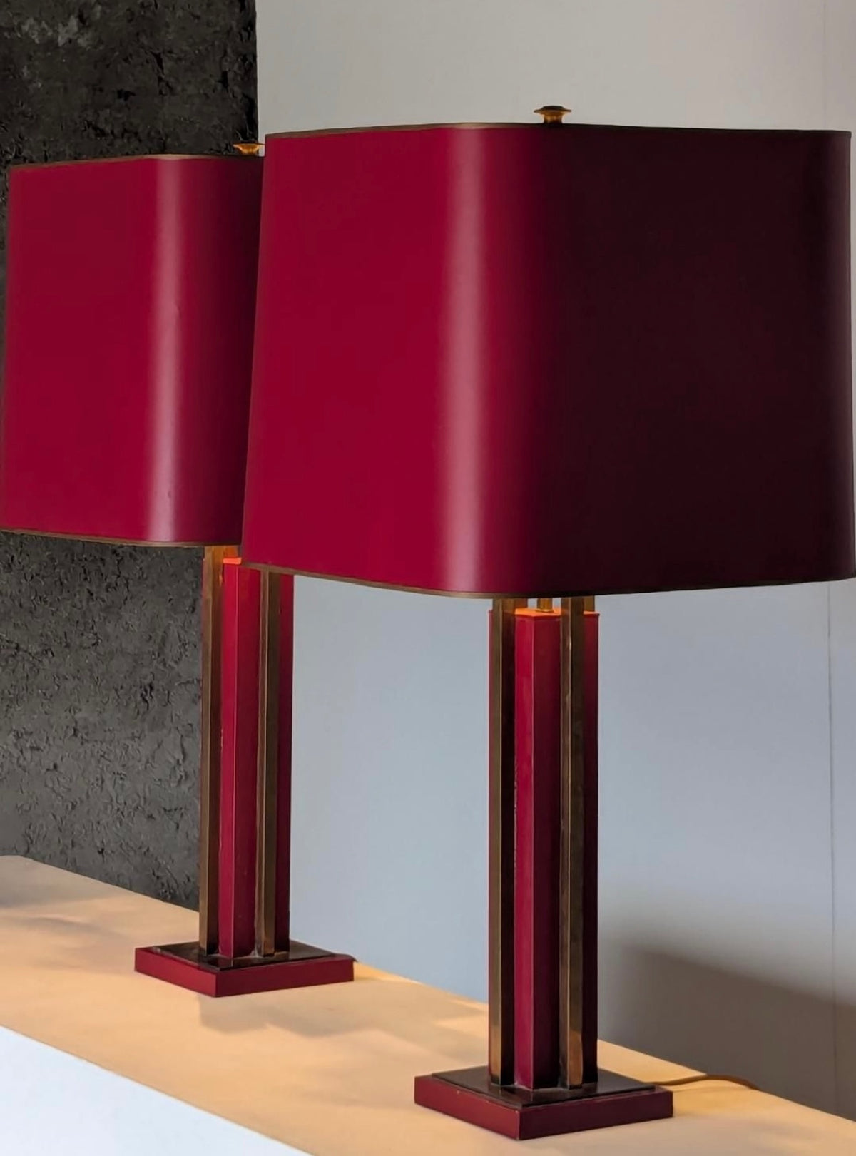 A pair of modern table lamps with large cylindrical deep red fabric shades and matching red and gold striped metal bases, displayed on a light-colored surface against a split gray and white background.