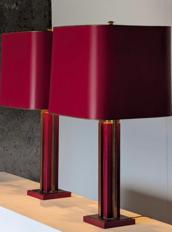 A pair of modern table lamps with large cylindrical deep red fabric shades and matching red and gold striped metal bases, displayed on a light-colored surface against a split gray and white background.