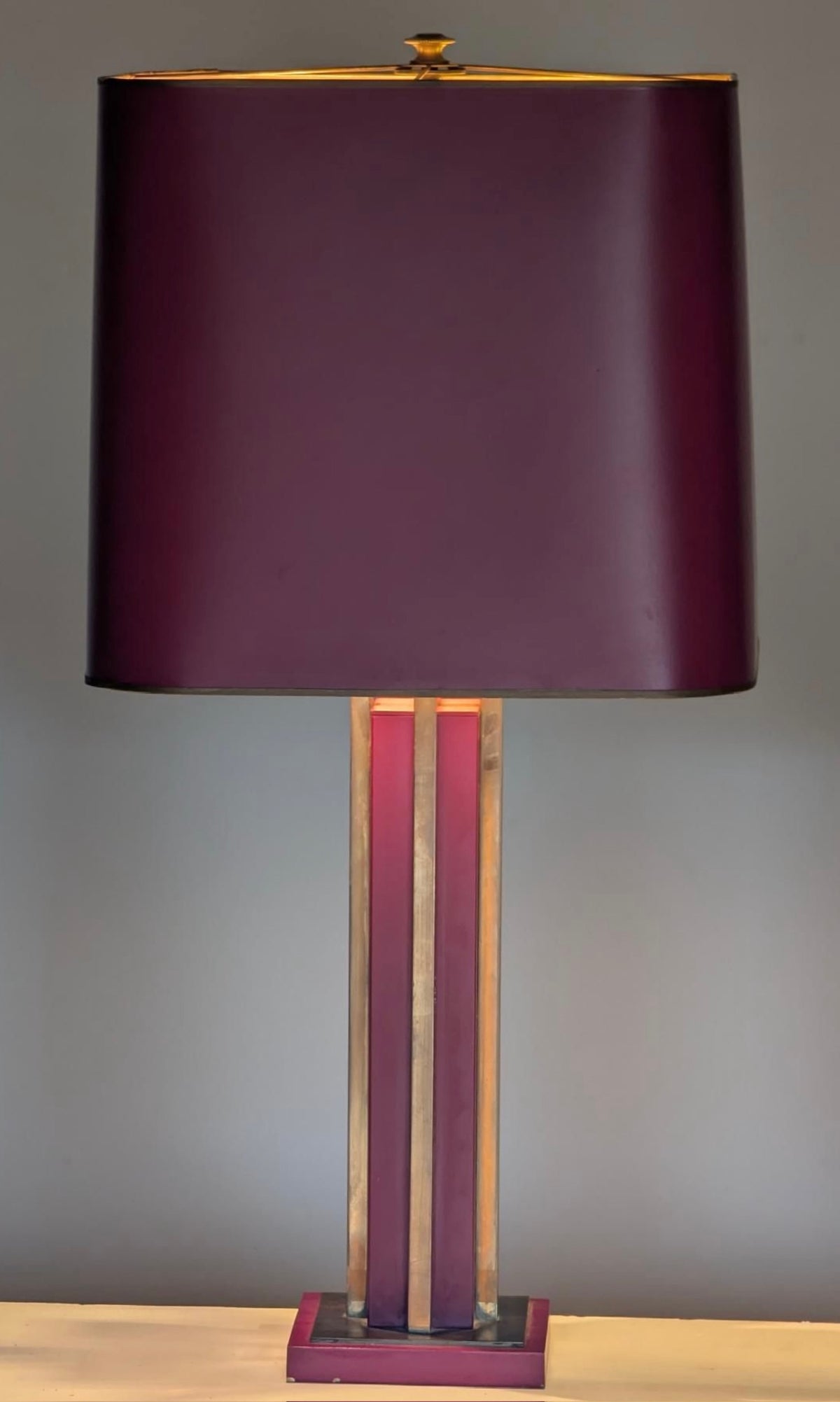 A table lamp with a large cylindrical deep burgundy shade, a matching burgundy base, and two vertical metallic gold-toned rods in the center, set against a plain gray background.
