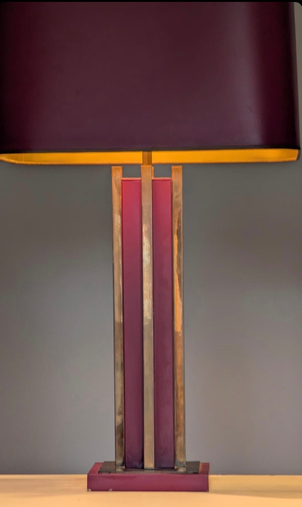 A table lamp with a large dark purple shade, a metallic gold inner rim, and a base featuring vertical panels of dark purple, bronze, and gold, set against a plain gray background.
