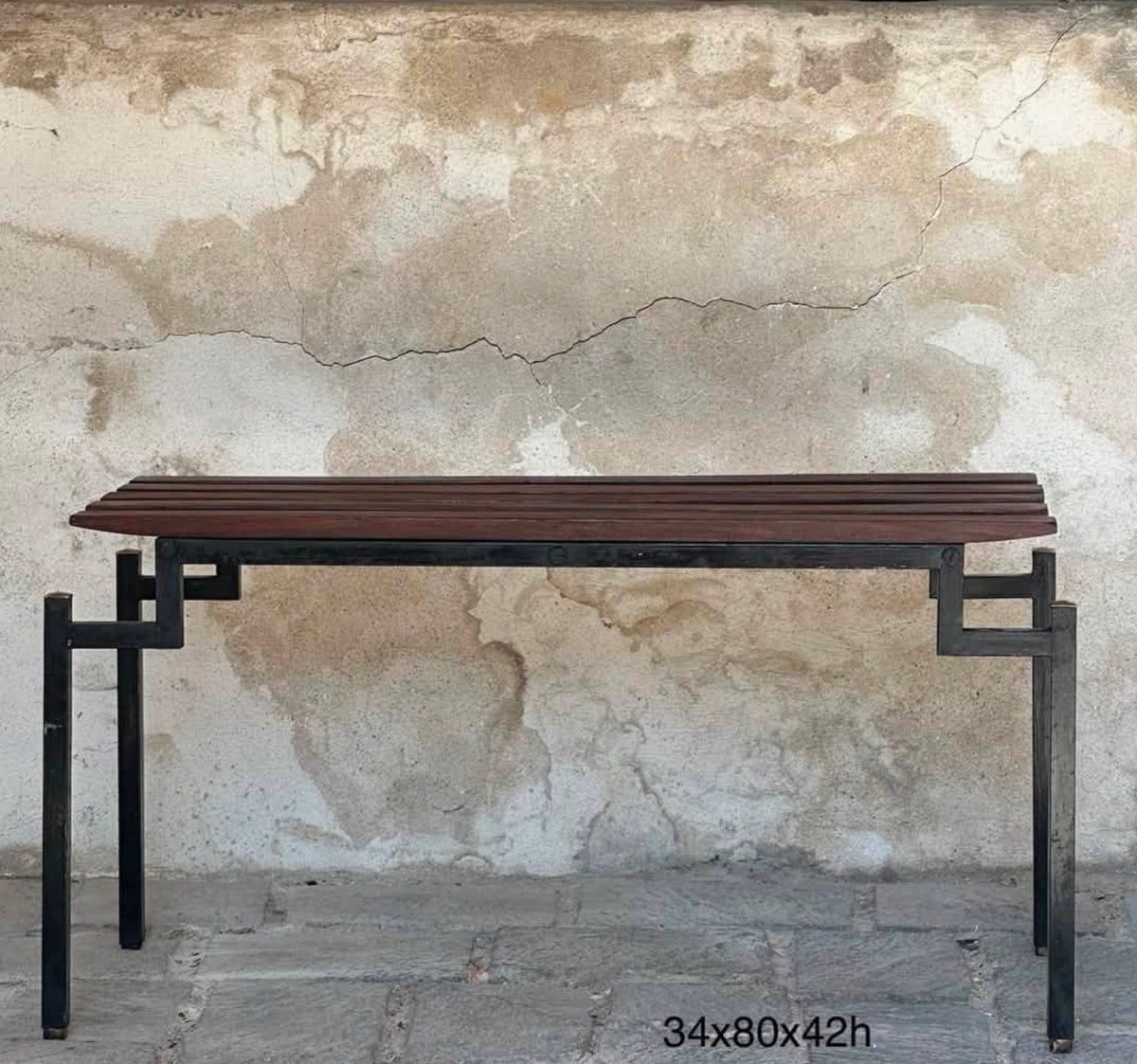A rectangular console table with a dark brown slatted top and a black metal frame, set against a textured, cracked plaster wall.