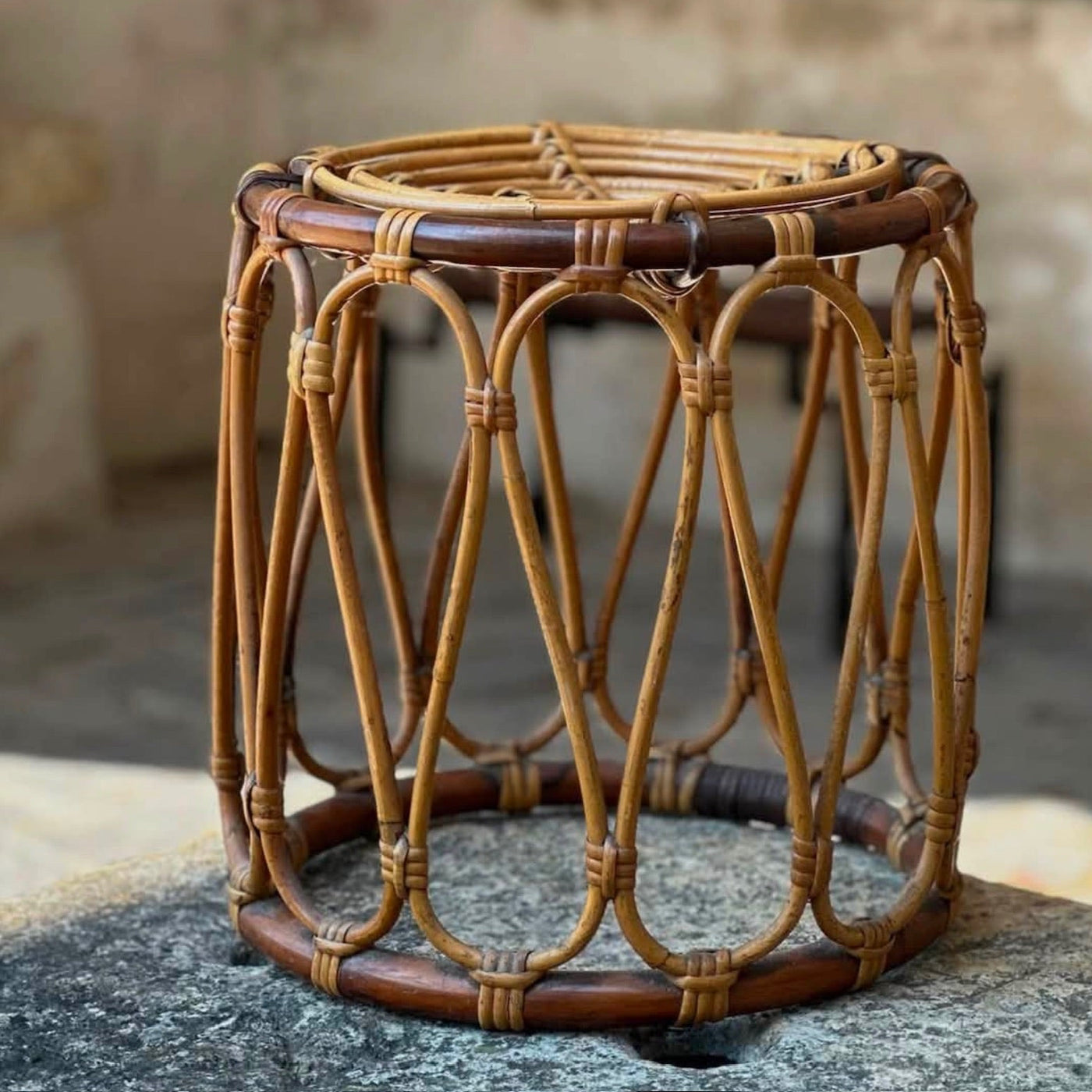 A round woven rattan stool with a natural tan finish and dark brown accents, shown on a stone surface in a rustic indoor setting.