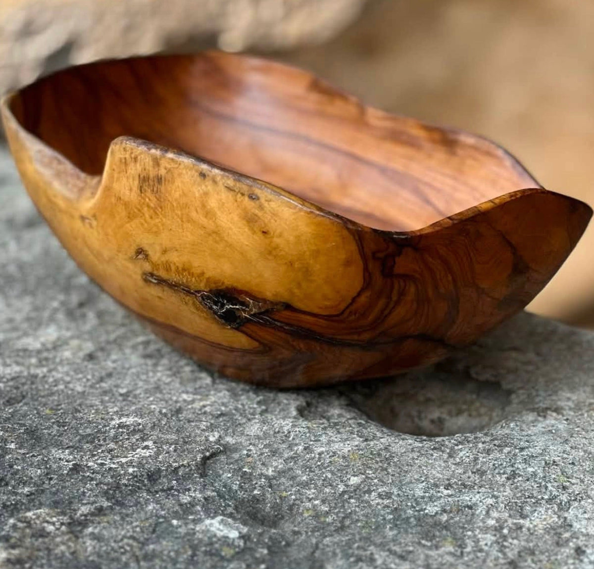 A handcrafted wooden bowl with organic, wavy edges, resting on a textured gray stone surface. The bowl features rich natural wood grain patterns in warm brown and golden-yellow tones.