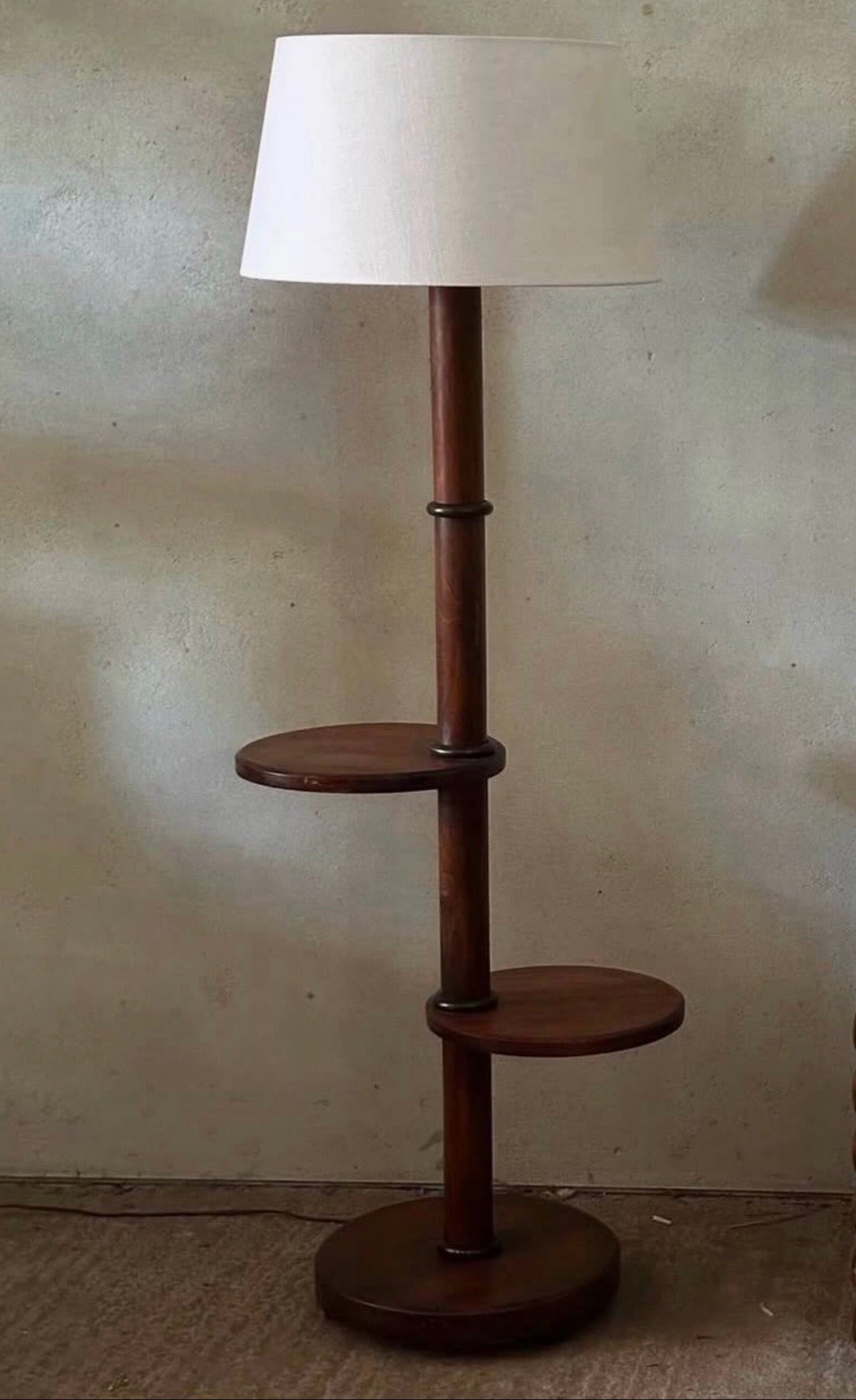 A mid-century modern floor lamp with a dark brown wooden pole, two circular shelves, and a large white drum shade, set against a textured beige wall.