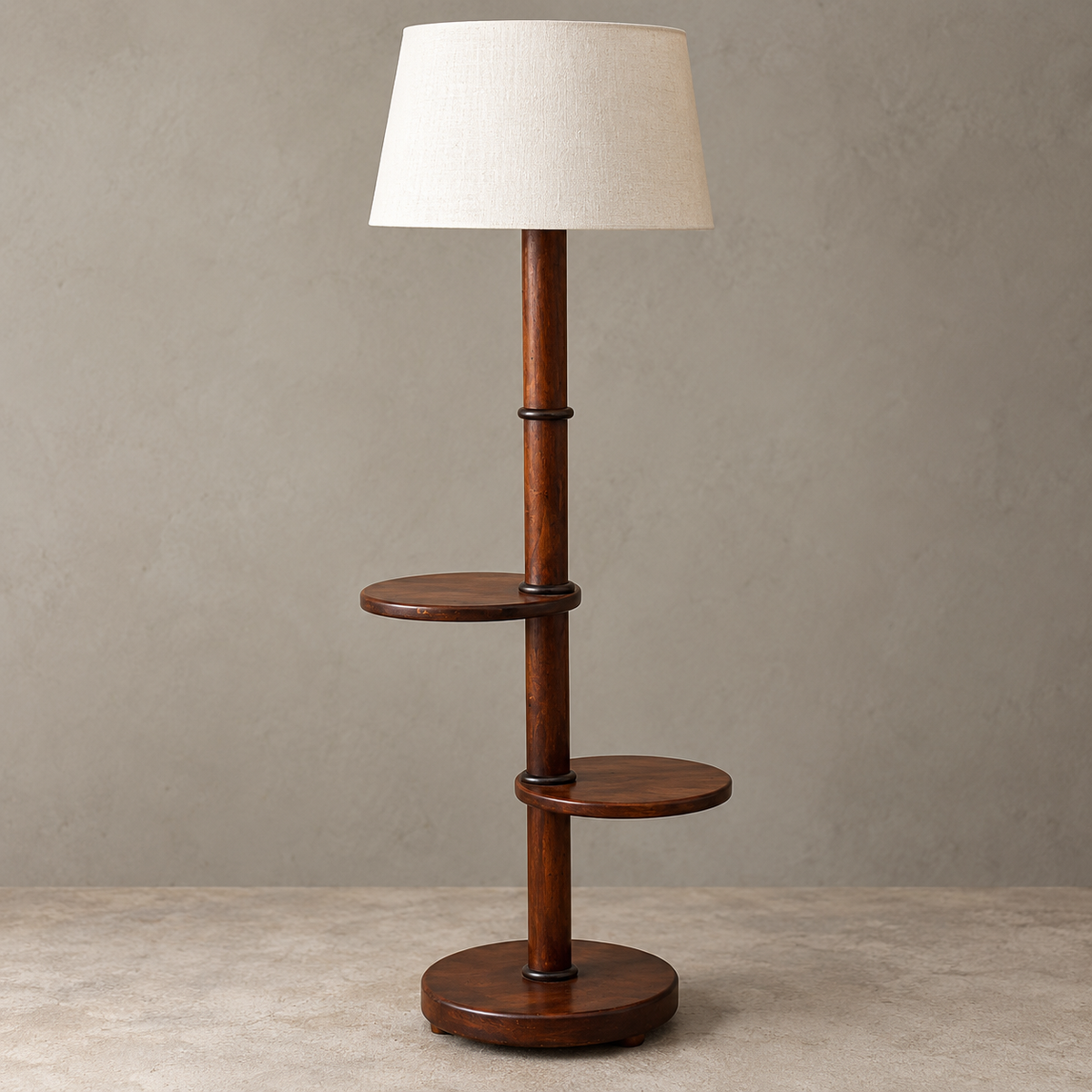 A modern wooden floor lamp with a tapered off-white fabric drum shade, a dark brown wood pole, two circular wooden shelves, and a round wooden base, set against a textured gray wall and concrete floor.