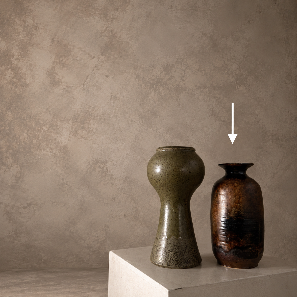 Two ceramic vases displayed on a white pedestal against a textured beige plaster wall. The vase on the left is olive green with a bulbous middle, and the vase on the right is dark brown with a flared rim, indicated by a white arrow pointing to it.