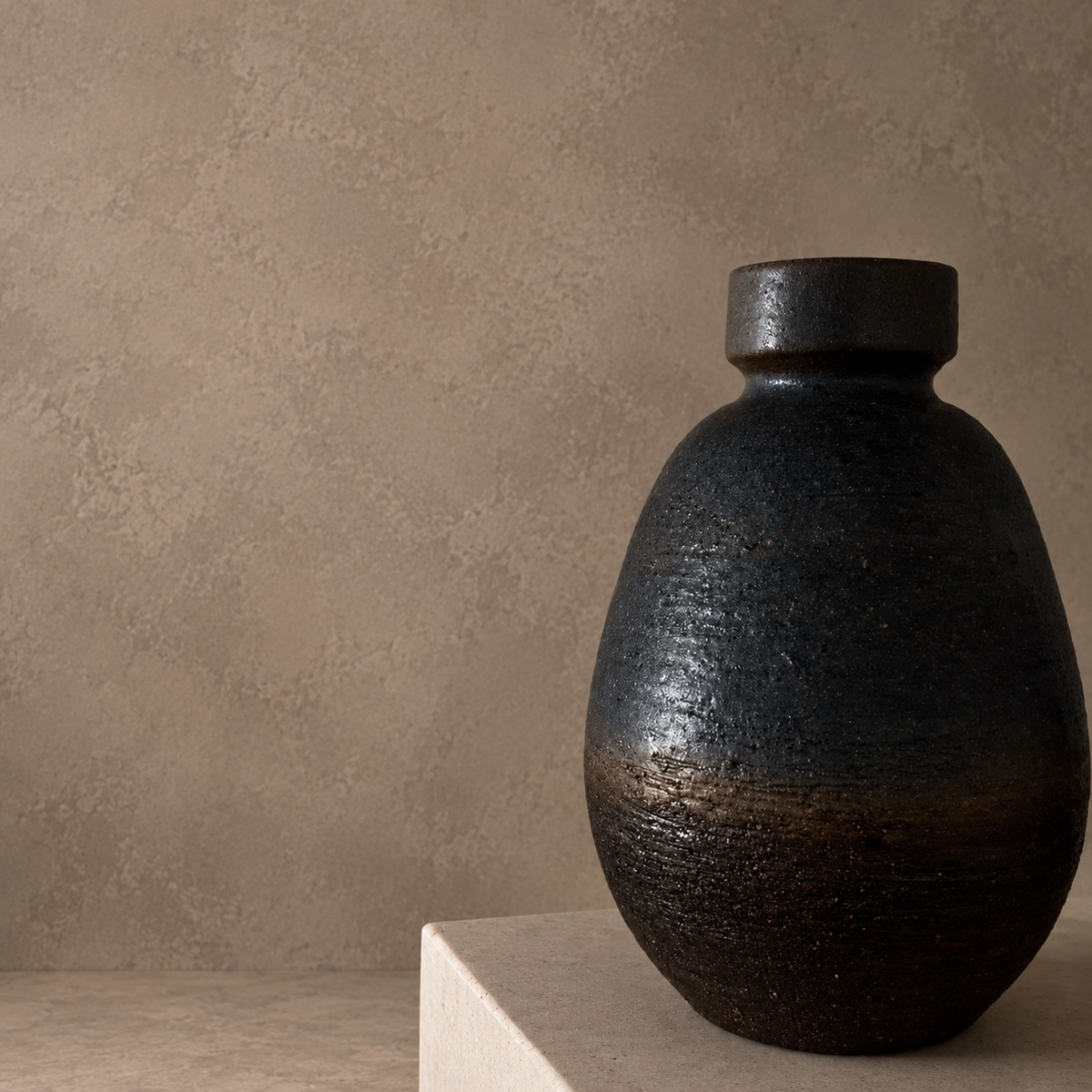 A dark glazed ceramic vase with a textured, metallic finish, resting on a light beige stone pedestal against a textured beige plaster wall.