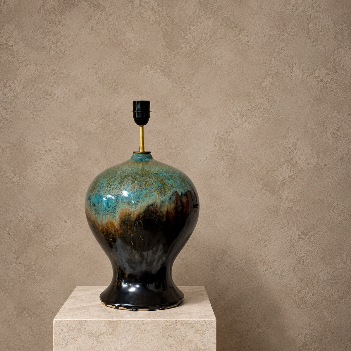 A ceramic table lamp with a glossy teardrop-shaped bulb in a turquoise, brown, and black glaze, featuring a gold metal stem and black socket, displayed on a stone pedestal against a textured beige wall.