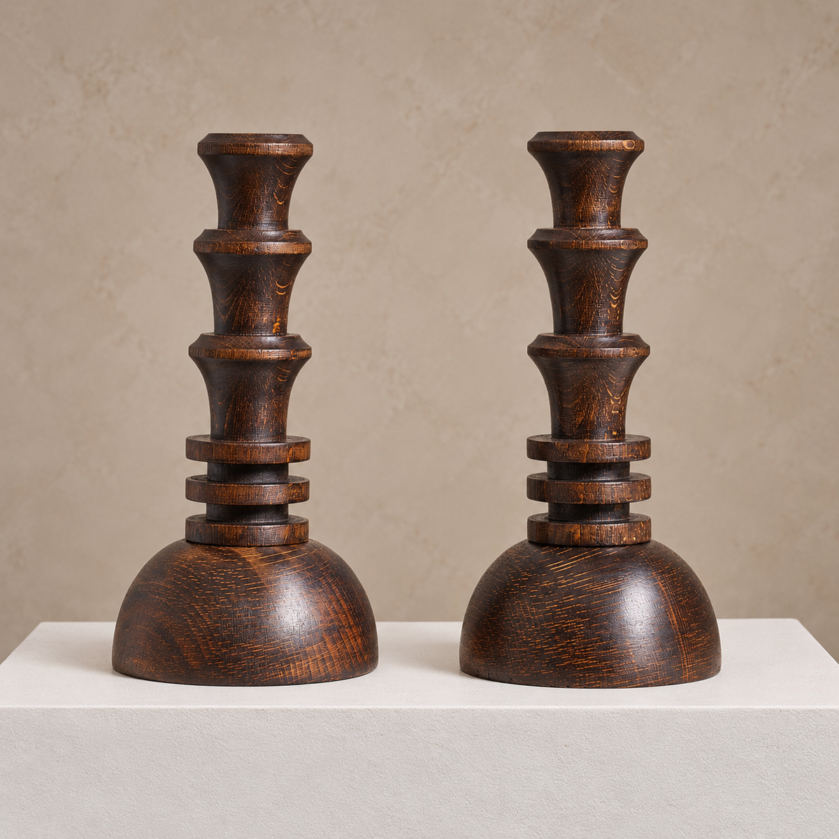 A pair of dark brown wooden decorative candlesticks with tiered stems and rounded bases, displayed on a white pedestal against a warm beige textured background.