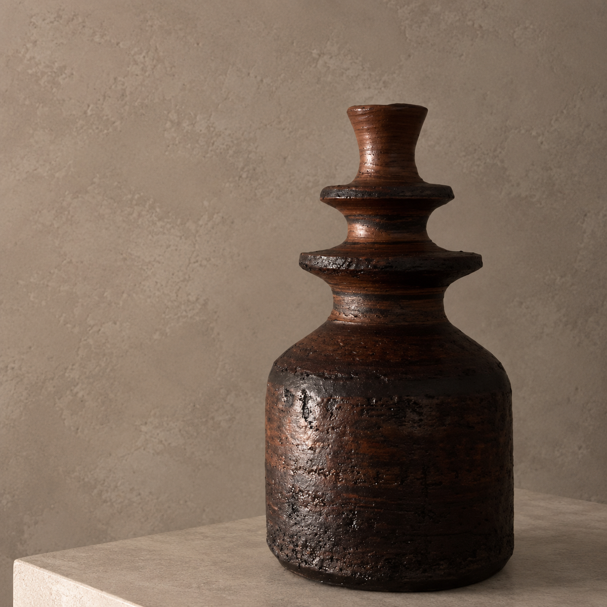 A rustic dark brown ceramic vase with a textured, weathered finish and a tiered neck, displayed on a light stone pedestal against a textured beige plaster wall.