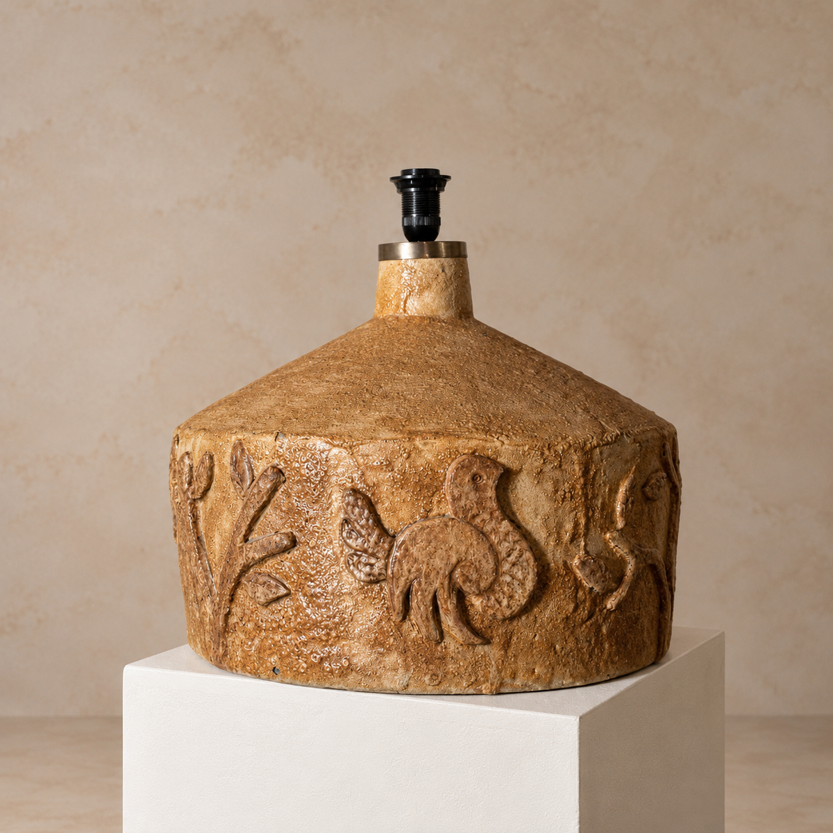 A ceramic lamp base with a textured, earthy brown body featuring carved floral and bird motifs, displayed on a white pedestal against a warm beige background.