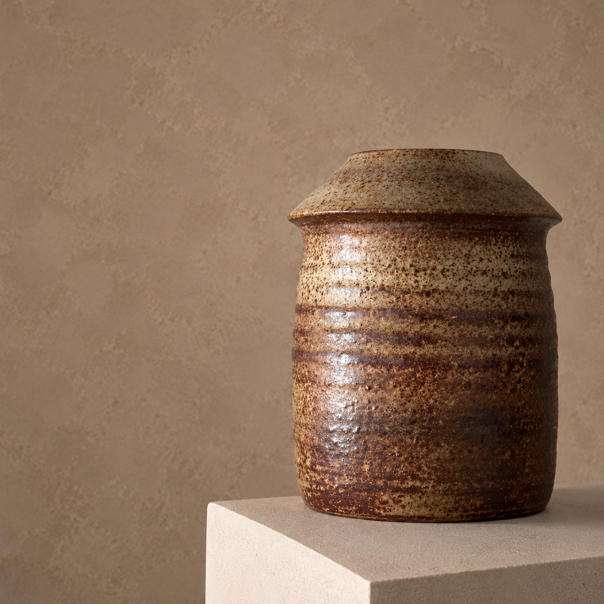 A rustic handmade ceramic jar with a textured, glazed finish in earthy brown and beige tones, displayed on a minimalist concrete pedestal against a textured beige wall.