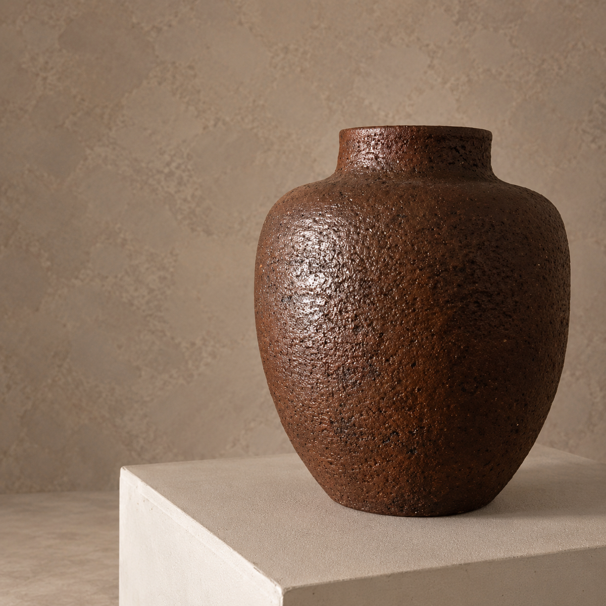 A textured brown ceramic vase with a rustic, speckled finish, displayed on a light beige pedestal against a neutral, textured beige wall.